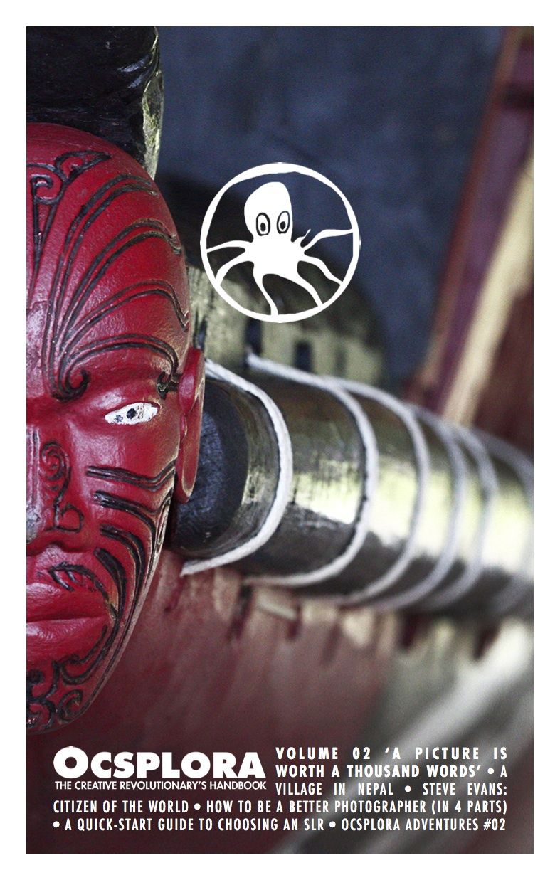 The cover of OCSPLORA Volume Two, carved wooden head at the front of a Maori longboat, red face with black tattoo lines and white eyes and black pupils